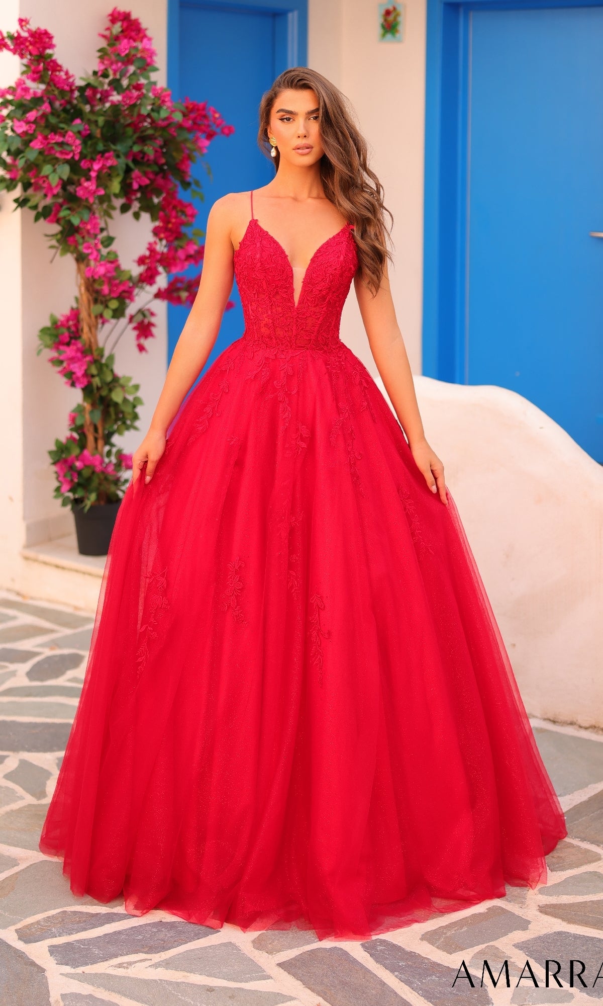 A woman stands outdoors on a stone path near blue doors and pink flowers, wearing the Amarra 88410 prom ball gown?a floor-length, sleeveless red dress with a deep V-neckline and corset bodice.